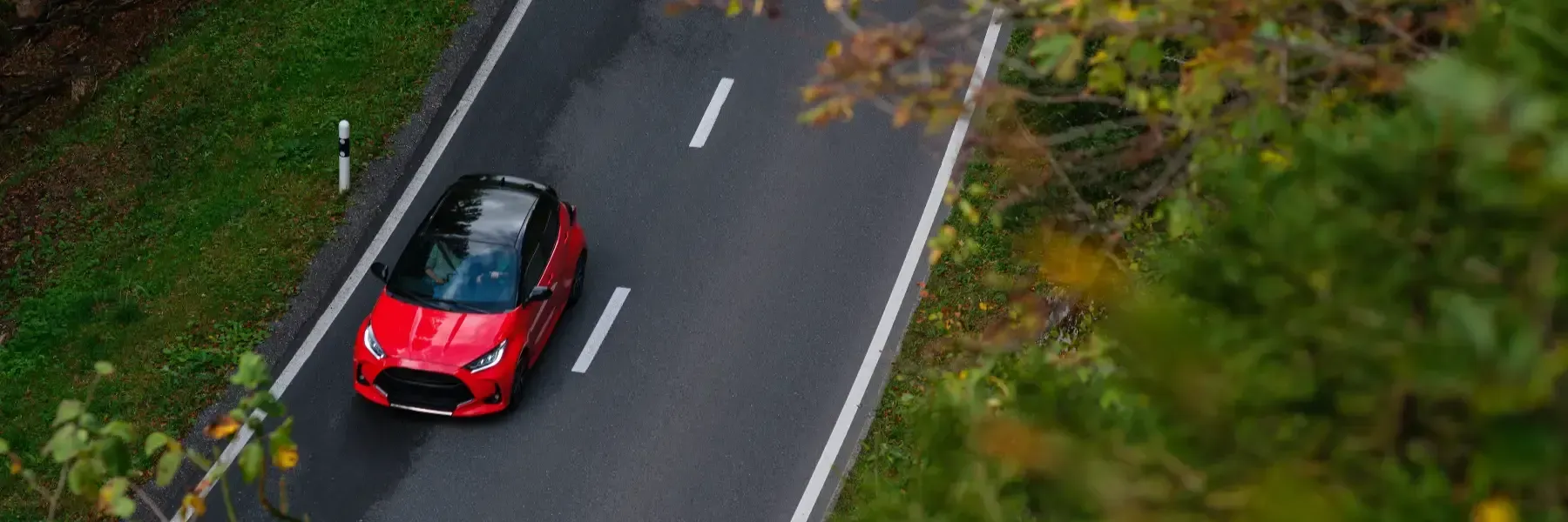 Vista aérea de un coche rojo con techo negro circulando por una carretera asfaltada bordeada de árboles.