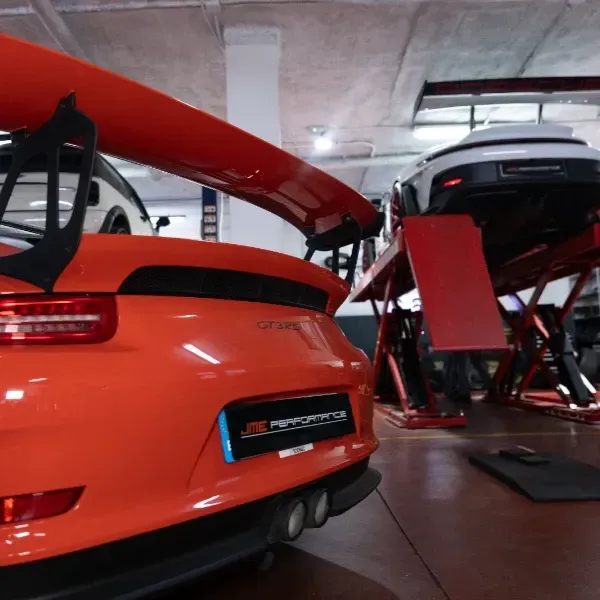 Coche deportivo naranja con alerón elevado en un garaje, visto desde atrás junto a otro coche en un elevador.