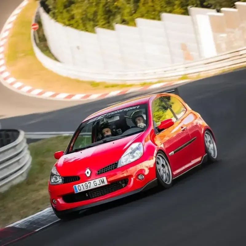 Un Renault Clio rojo circulando por un circuito, tomando una curva con dos personas en su interior.