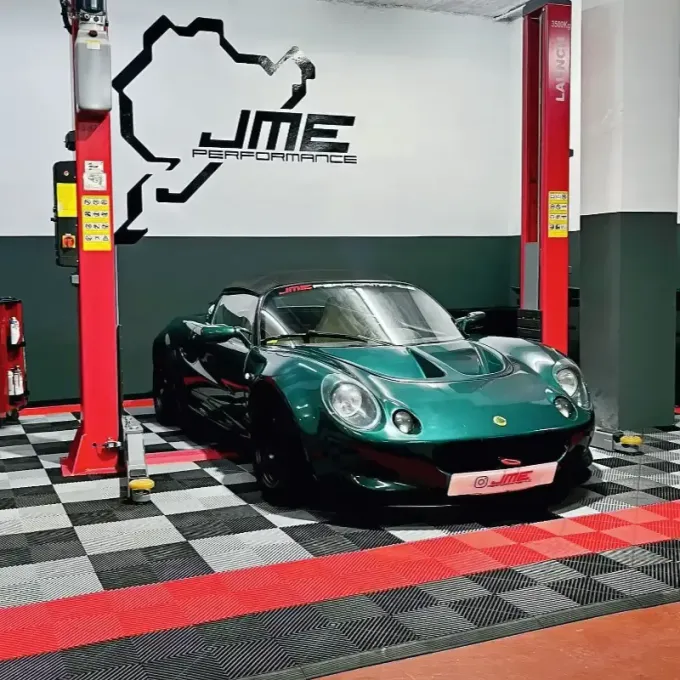 Un deportivo Lotus de color verde oscuro aparcado en un garaje con columnas de elevación rojas y suelo a cuadros.