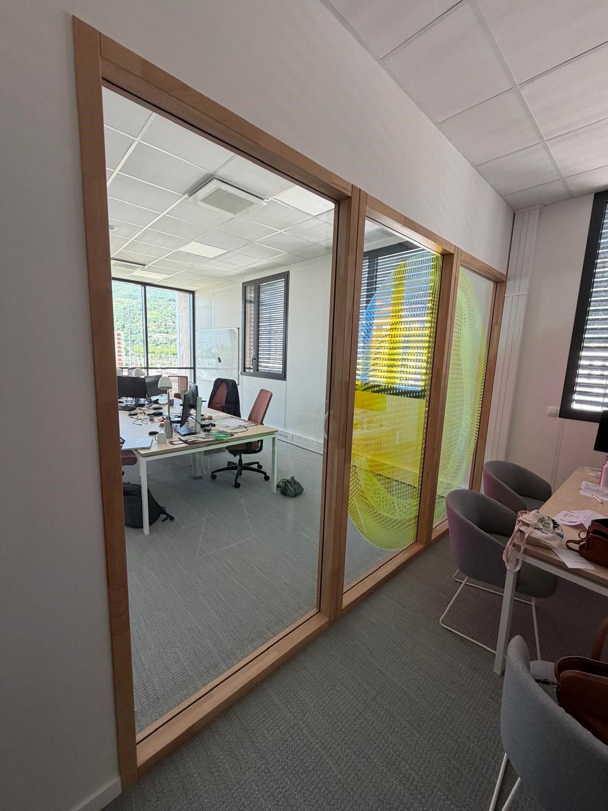 Couloir de bureaux donnant sur une grande salle de réunion vitrée, avec des bureaux visibles à l'intérieur.