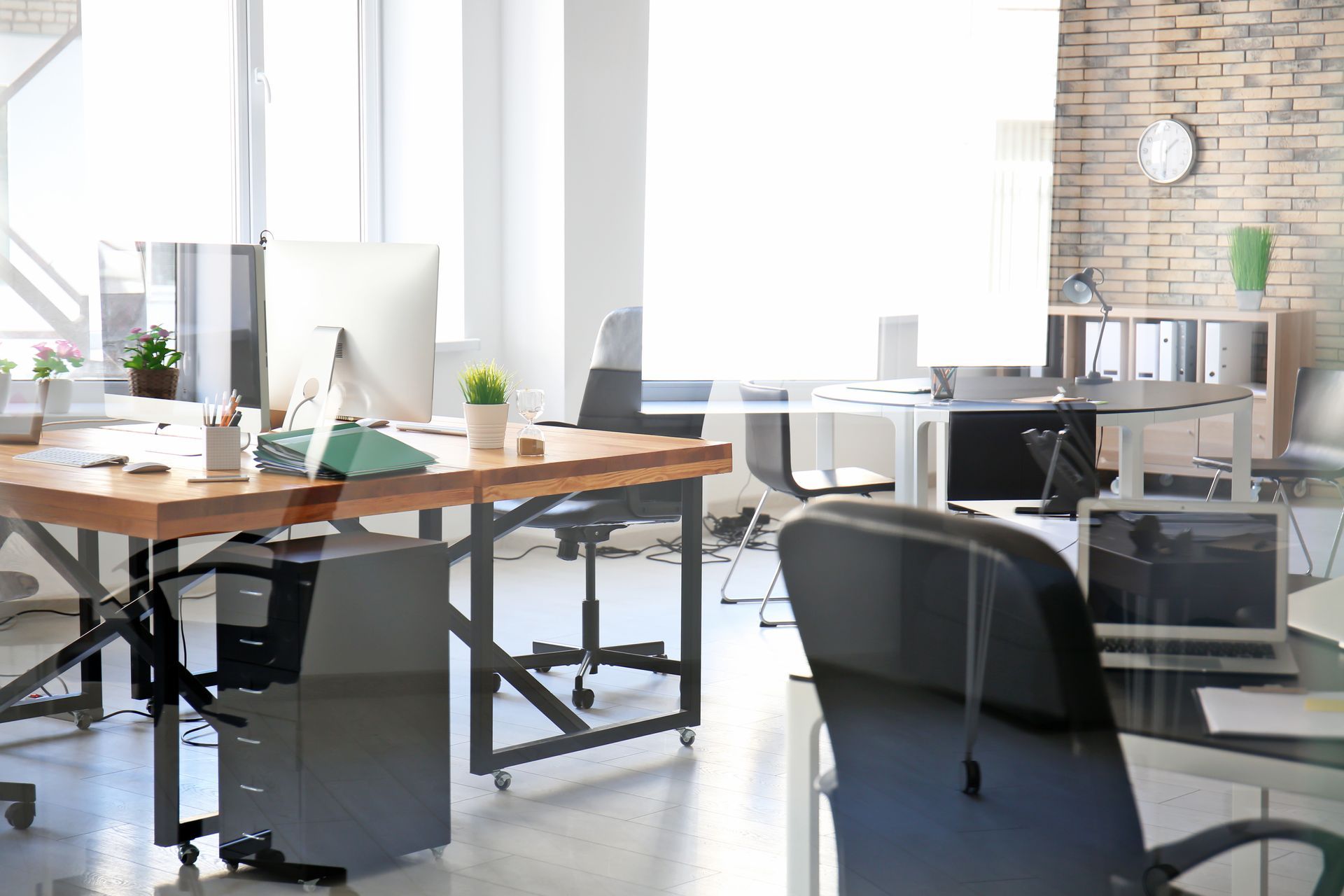 Bureau moderne et lumineux avec bureaux en bois, ordinateurs, chaises noires et grandes fenêtres.