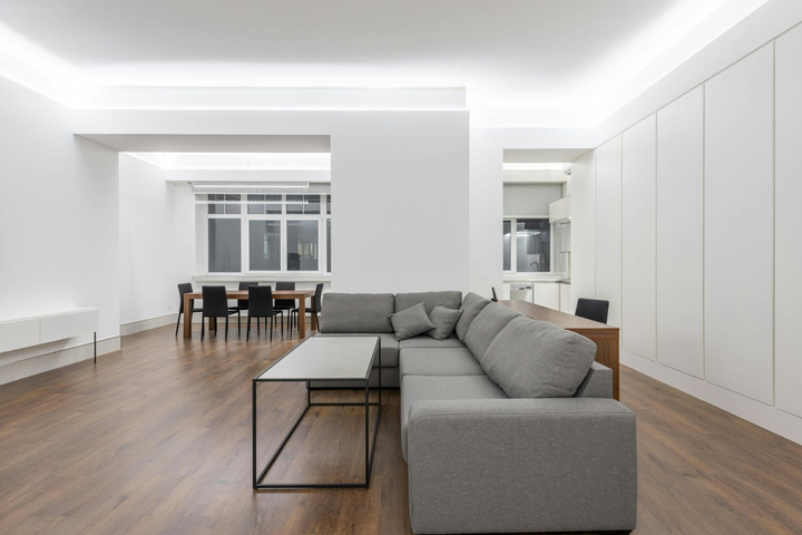 Salon moderne et lumineux avec canapé d'angle gris, parquet et coin repas en arrière-plan.