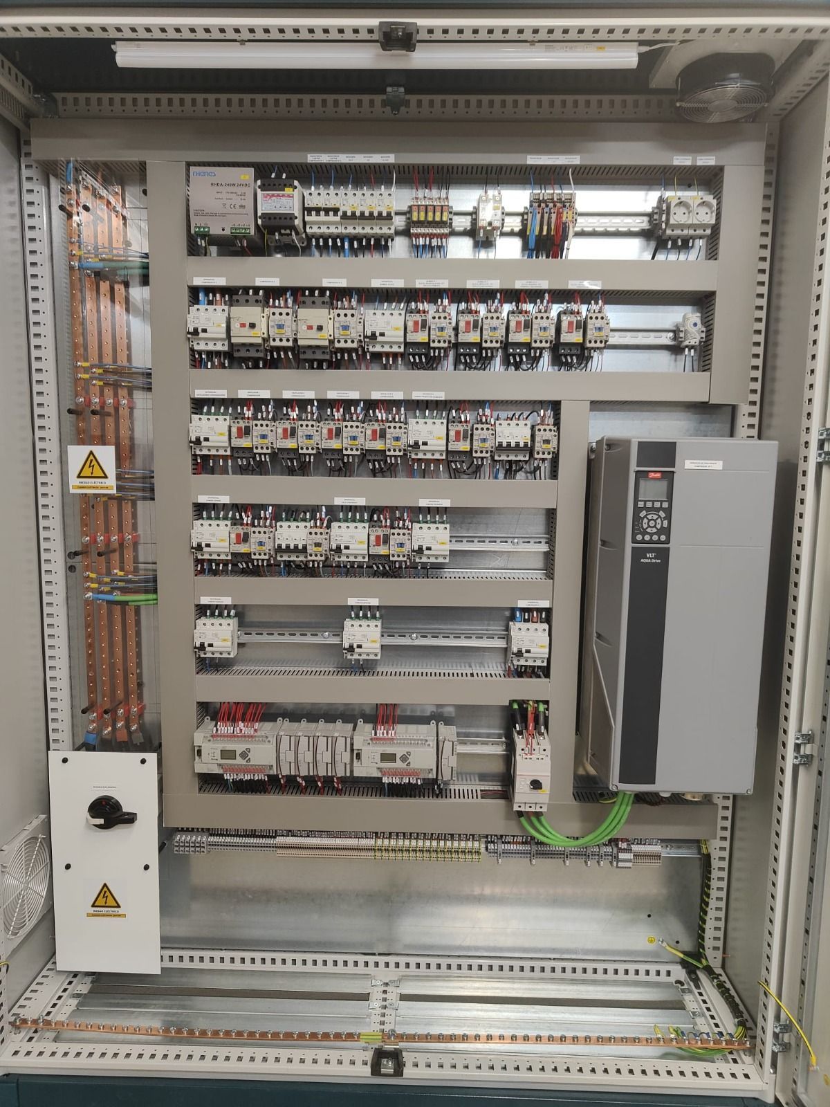 Un panel de control eléctrico abierto con filas de disyuntores, contactores y una gran unidad de accionamiento de color gris.