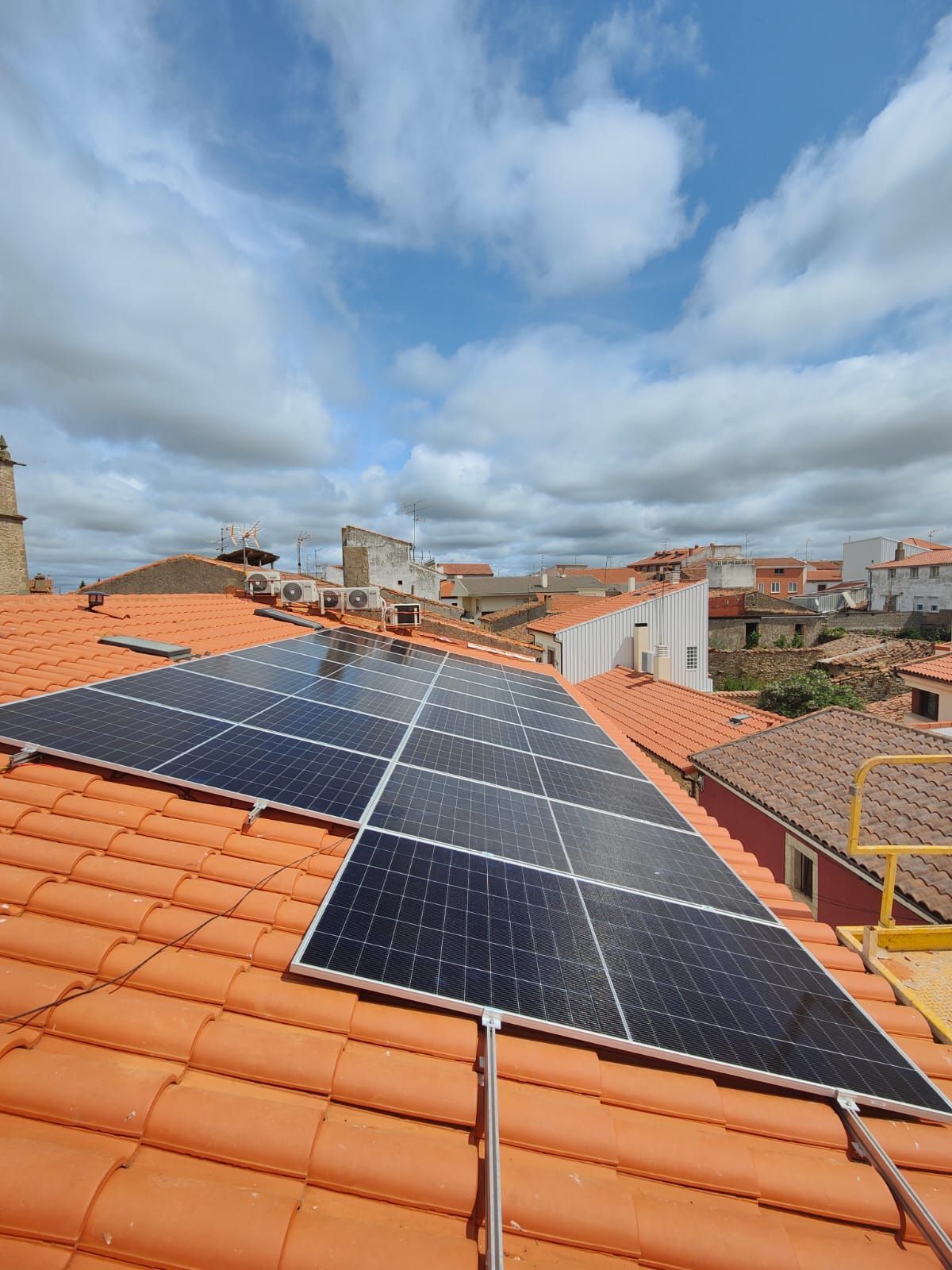 Paneles solares instalados en un tejado de tejas naranjas bajo un cielo azul nublado en un barrio residencial.