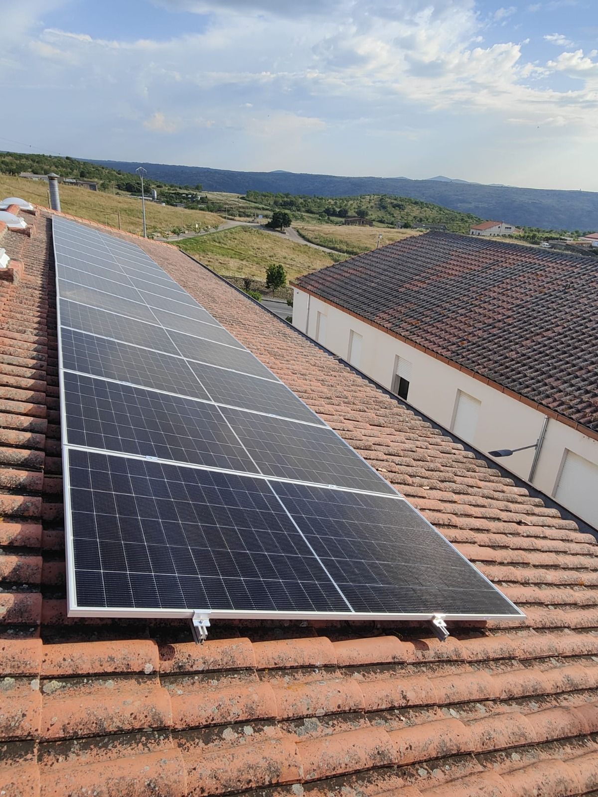 Un conjunto rectangular de paneles solares negros montados sobre un tejado de tejas rojas, con vistas a un paisaje rural.