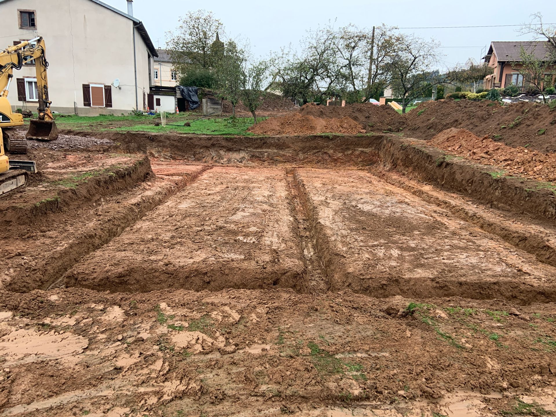 Chantier de construction excavé, avec de la terre boueuse, des tranchées de fondation nivelées et des maisons en arrière-plan