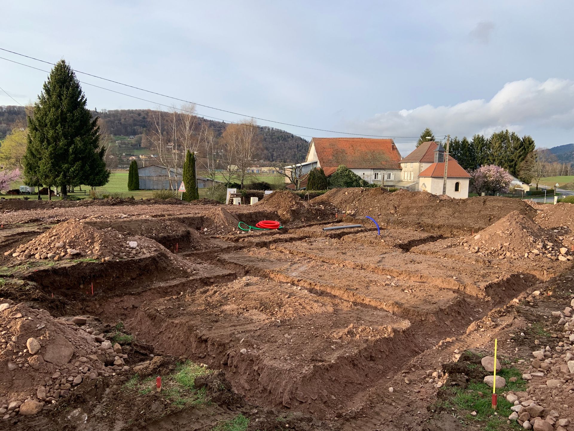 Terrain en friche après excavation, avec d’importants travaux de terrassement, des maisons aux toits rouges et une colline en arrière-plan sous un ciel nuageux
