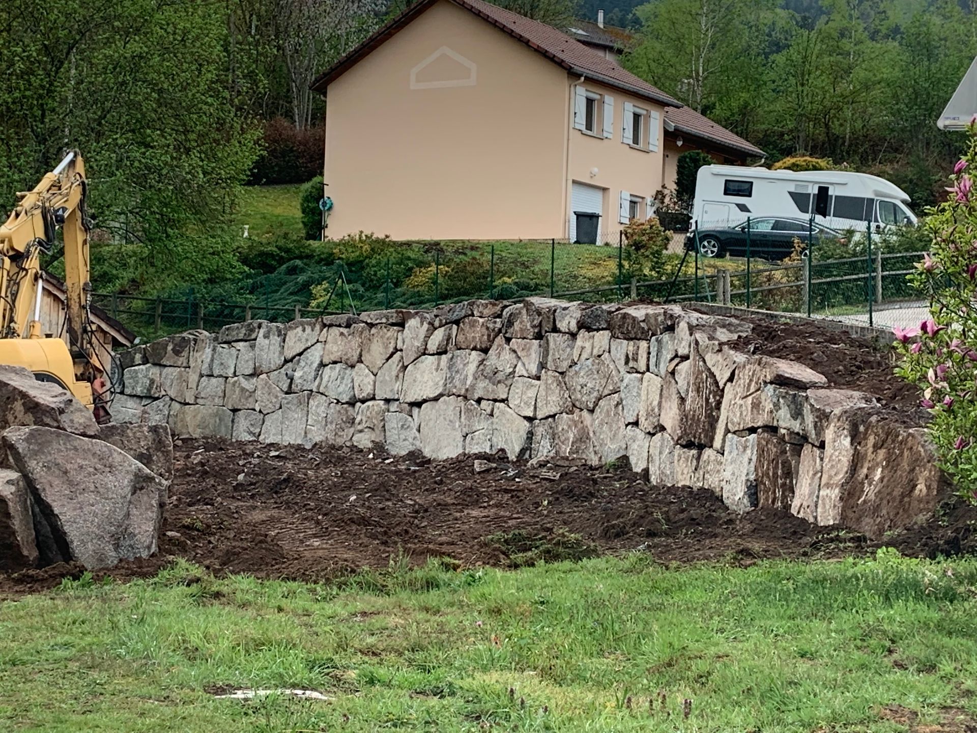 Mur de soutènement en pierre en cours de construction à côté d’une maison, avec une excavatrice et des travaux de terrassement dans un jardin herbeux