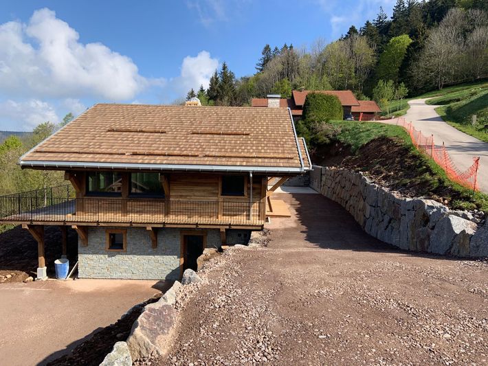Maison rustique avec un rez-de-chaussée en pierre et un toit en bois, au bord d’une route à flanc de colline sous un ciel bleu