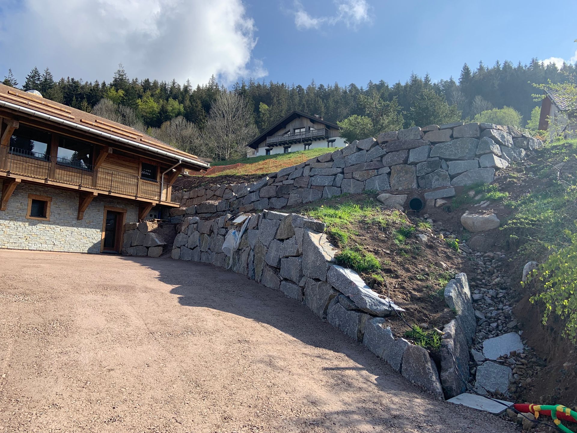 Mur de soutènement en pierre près d’une maison rustique en bois, sur un coteau ensoleillé sous un ciel bleu.