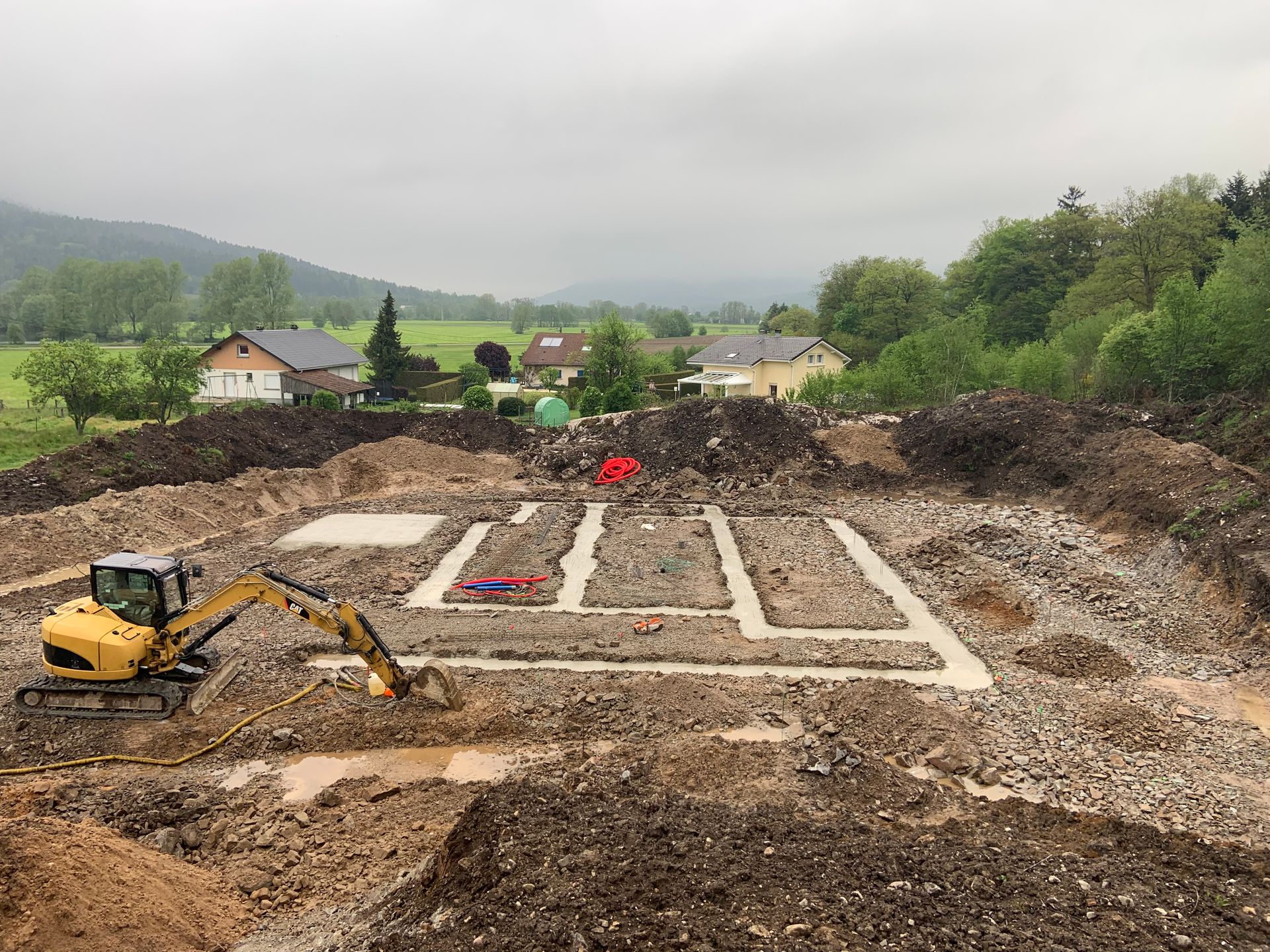 Une pelleteuse sur un chantier, avec des tranchées de fondation et des ouvriers sur un terrain boueux.