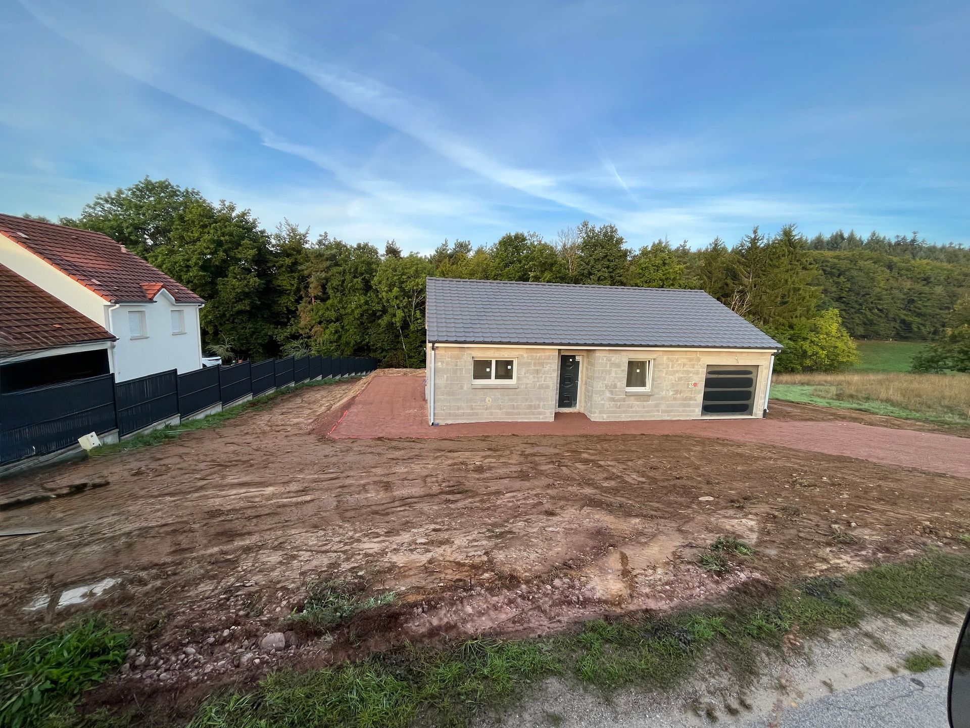 Petite maison inachevée au toit gris, sur un terrain défriché et terreux, près d’une route et d’arbres