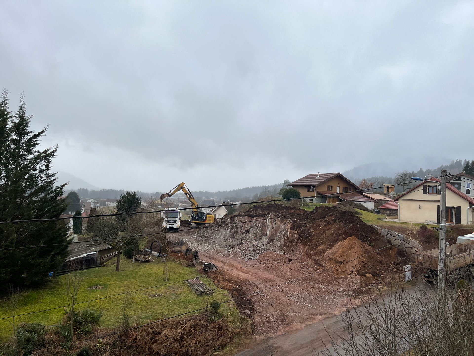 Une excavatrice dégage un important terrassement de terrain près de maisons, dans un quartier rural, sous un ciel couvert