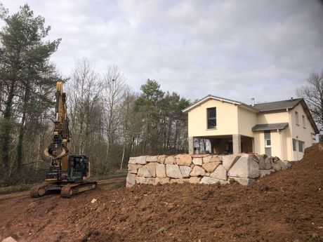 Une pelleteuse nivelle un terrain en terre à côté d’une maison beige, avec un mur de soutènement en pierre et des arbres en arrière-plan