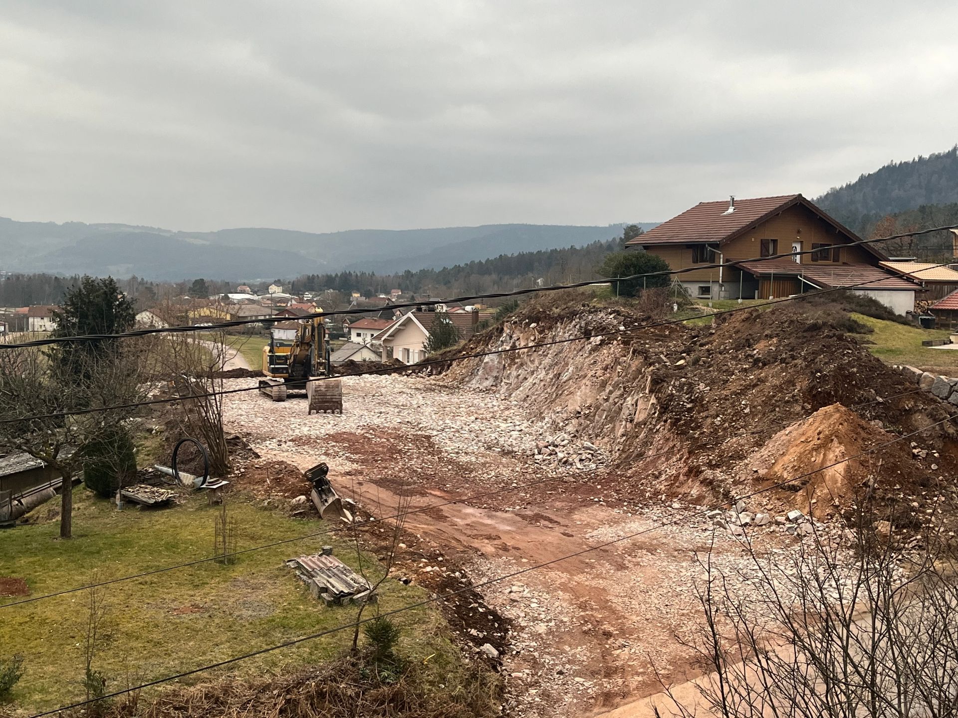 Un terrassement de terrain, traverse un coteau rural près de maisons, sous un ciel couvert.