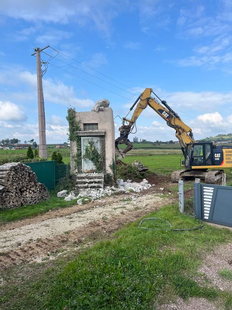 Une pelleteuse démolit une structure en béton au bord d’un chemin de terre, dans un champ à la campagne