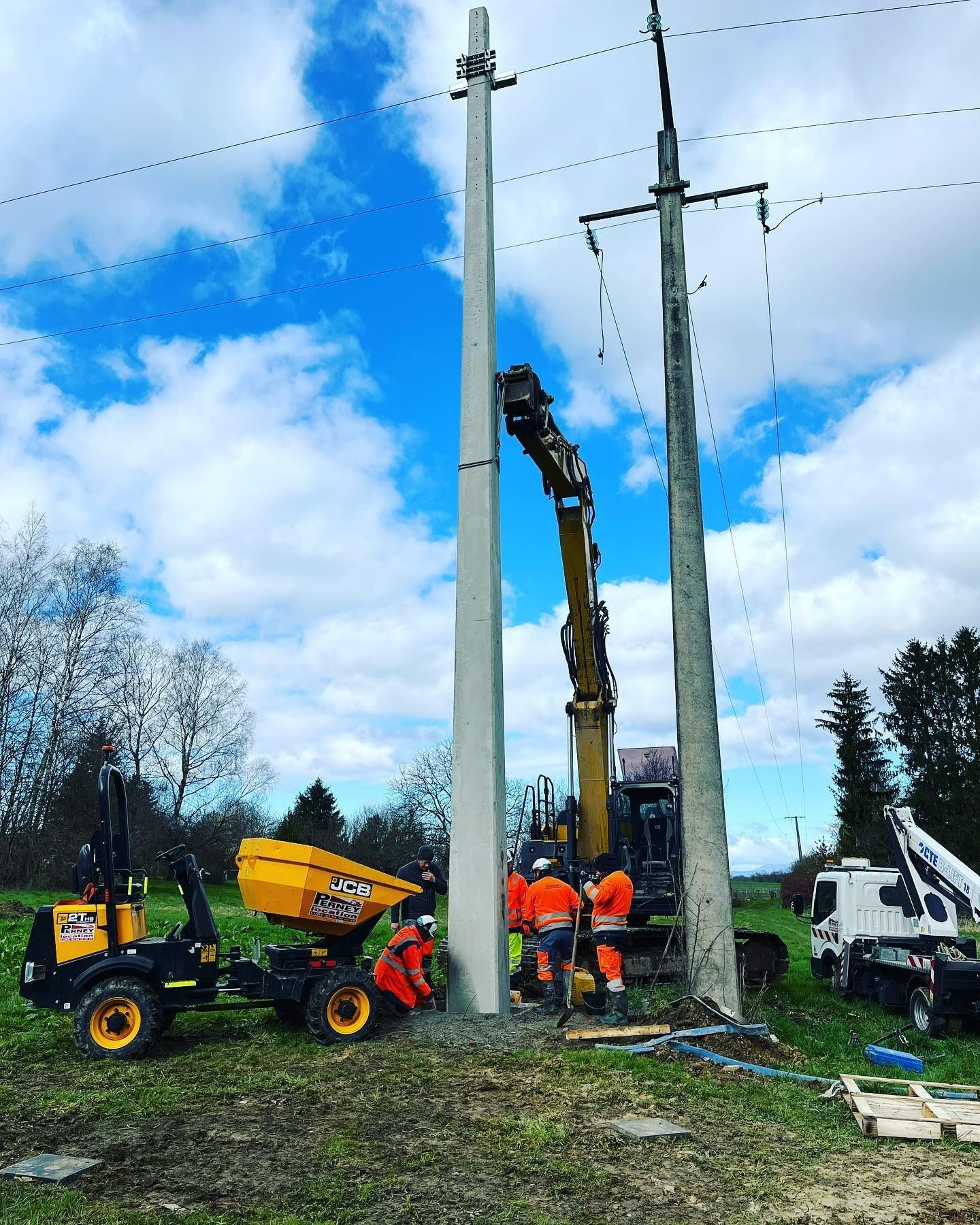 Des employés des services publics avec un engin de chantier réparent un poteau électrique en bordure de route, sous un ciel bleu.