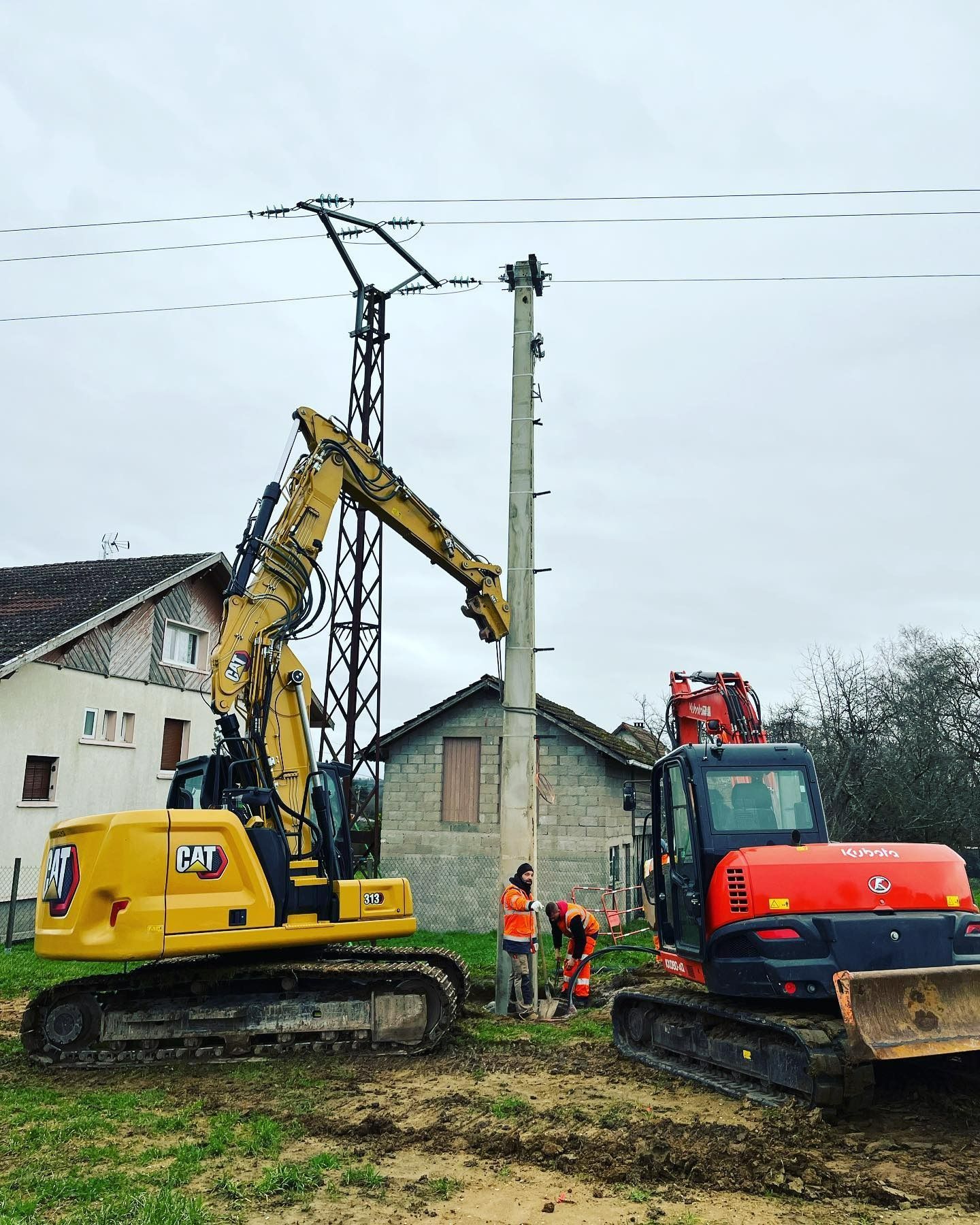 Des engins de chantier soulèvent un poteau électrique en béton à côté d'une maison en bordure de route rurale.