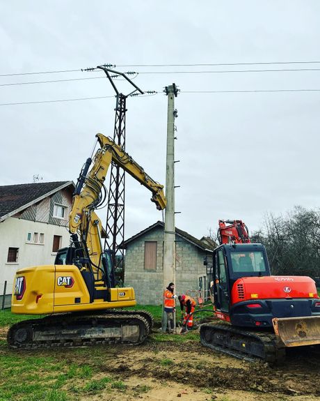 Des engins de chantier soulèvent un poteau électrique en béton à côté d'une maison en bordure de route rurale.