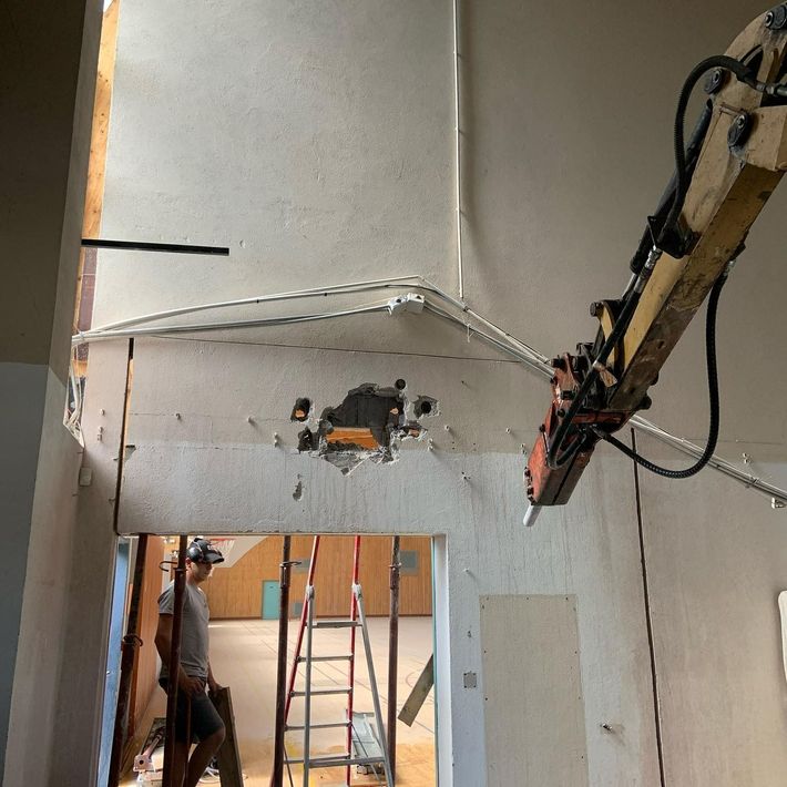 Cloison intérieure partiellement construite avec plaques de plâtre apparentes, une échelle et un ouvrier utilisant un outil à long manche.