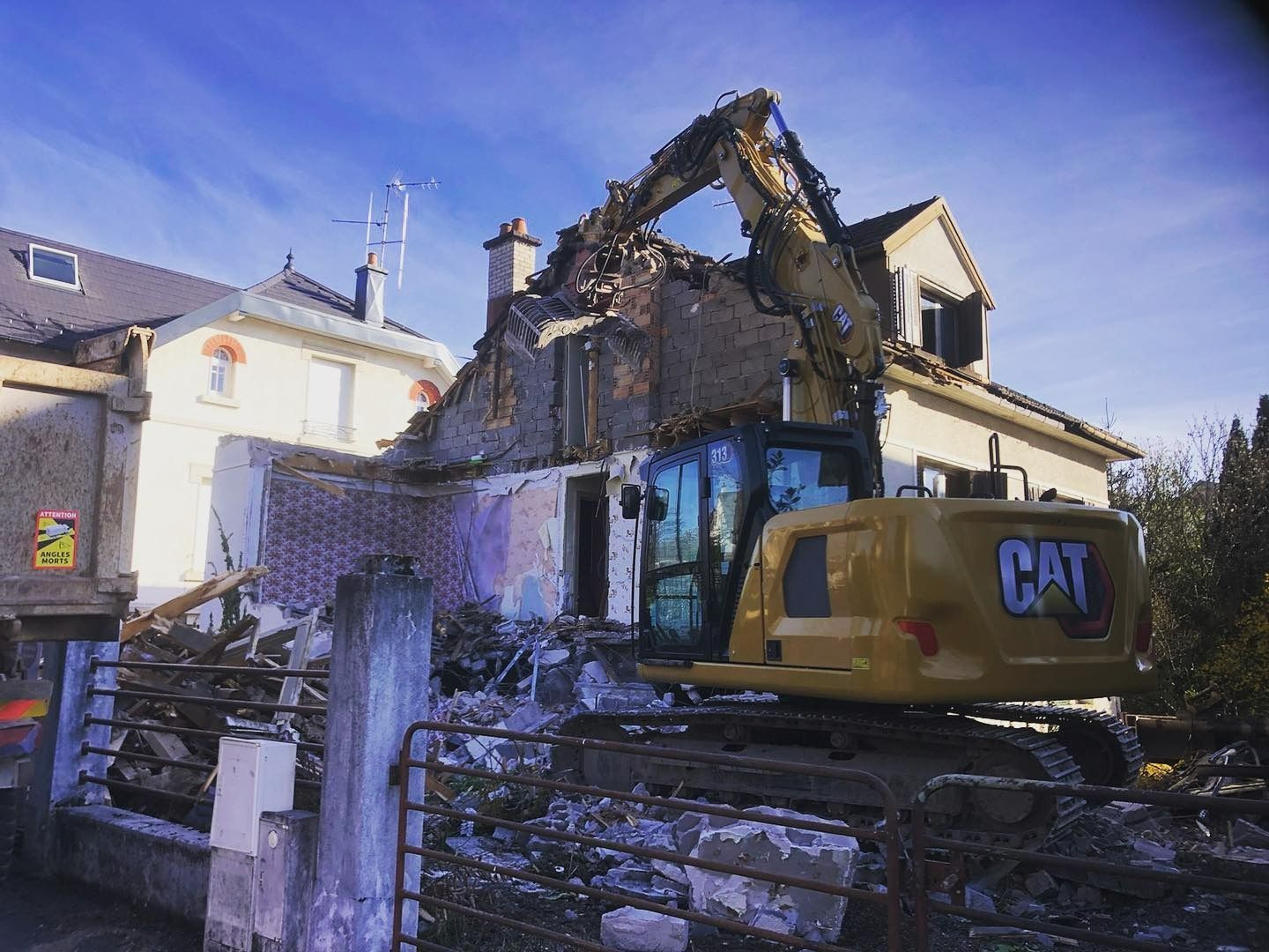 Maison endommagée avec un toit effondré à côté d'une pelleteuse CAT jaune et de décombres sous un ciel bleu.