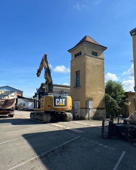 Une pelleteuse CAT jaune à côté d'une haute tour en pierre sur un terrain gravillonné sous un ciel bleu.