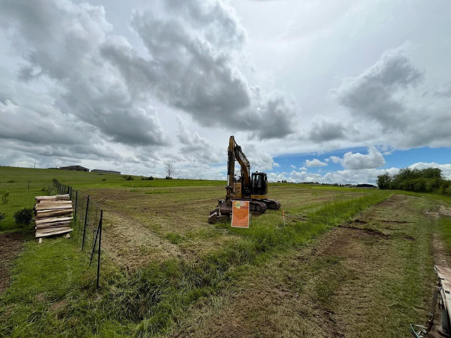 Une excavatrice travaille dans un champ herbeux sous un ciel nuageux, avec des planches empilées à gauche.
