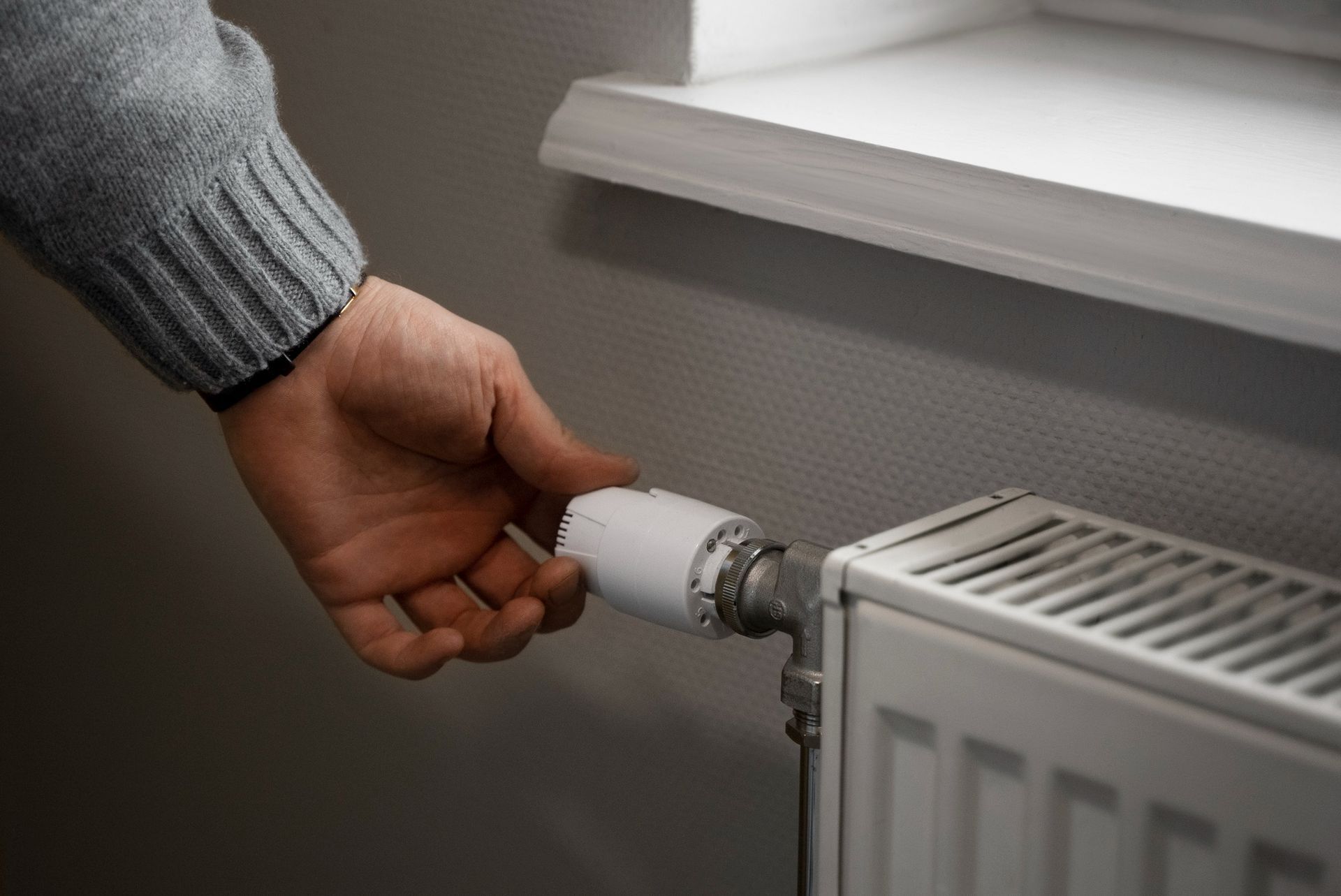 La mano de una persona girando la perilla blanca de un termostato en un radiador cerca de una ventana.