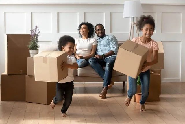 Famille dans une nouvelle maison avec des enfants portant des cartons, des parents souriants sur un canapé.