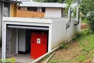 Auf einem grasbewachsenen Hang befindet sich ein Generator in einem offenen, weißen Betongehäuse mit zwei Abluftöffnungen.