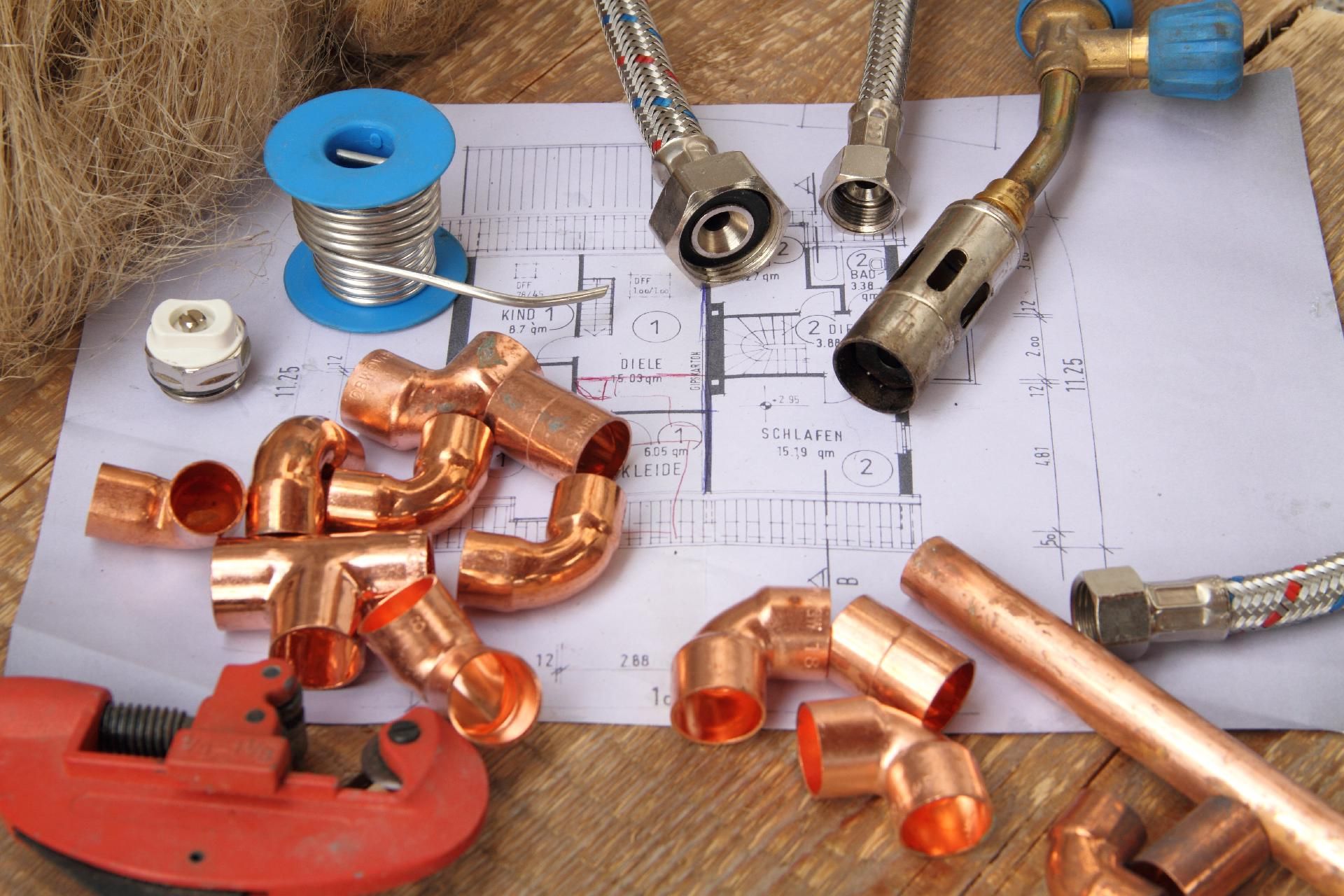 Kupferne Sanitärarmaturen, Rohrstücke und Werkzeuge auf einer mit Bauplänen bedeckten Werkbank