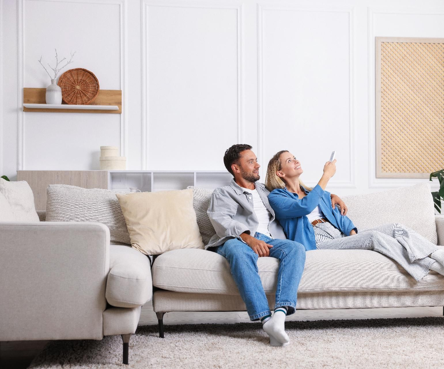 Ein Paar sitzt auf einem Sofa in einem Wohnzimmer und zeigt mit einer Fernbedienung auf etwas außerhalb des Bildausschnitts.