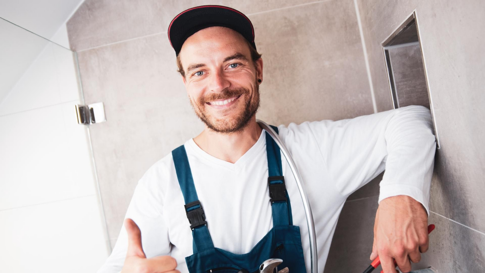 Ein lächelnder Arbeiter in blauem Overall und roter Mütze lehnt in einem gefliesten Badezimmer.