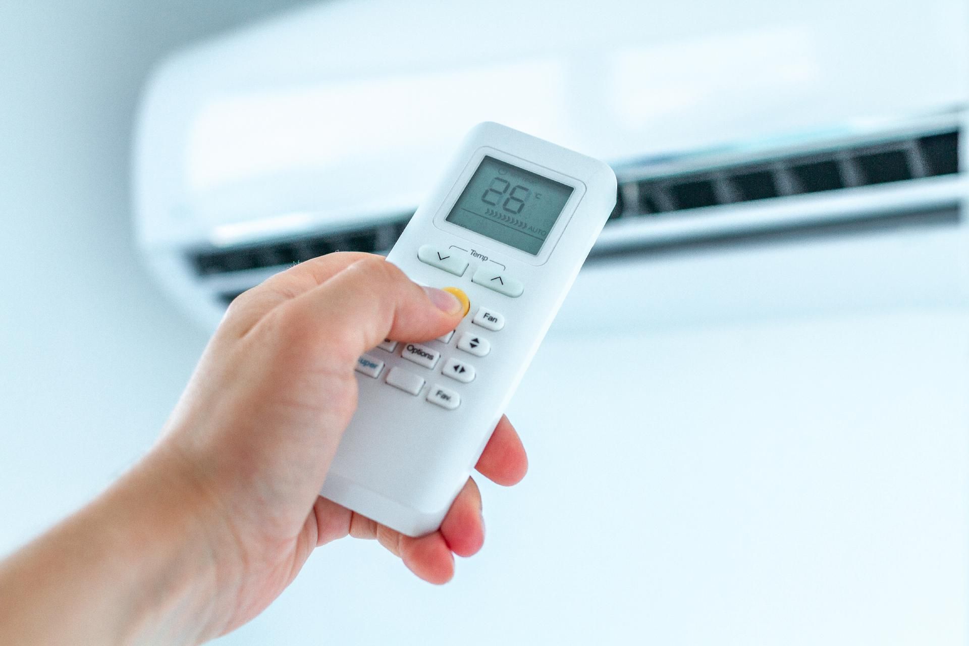 Eine Hand hält eine weiße Fernbedienung für eine Klimaanlage und richtet sie auf ein an der Wand montiertes Klimagerät.