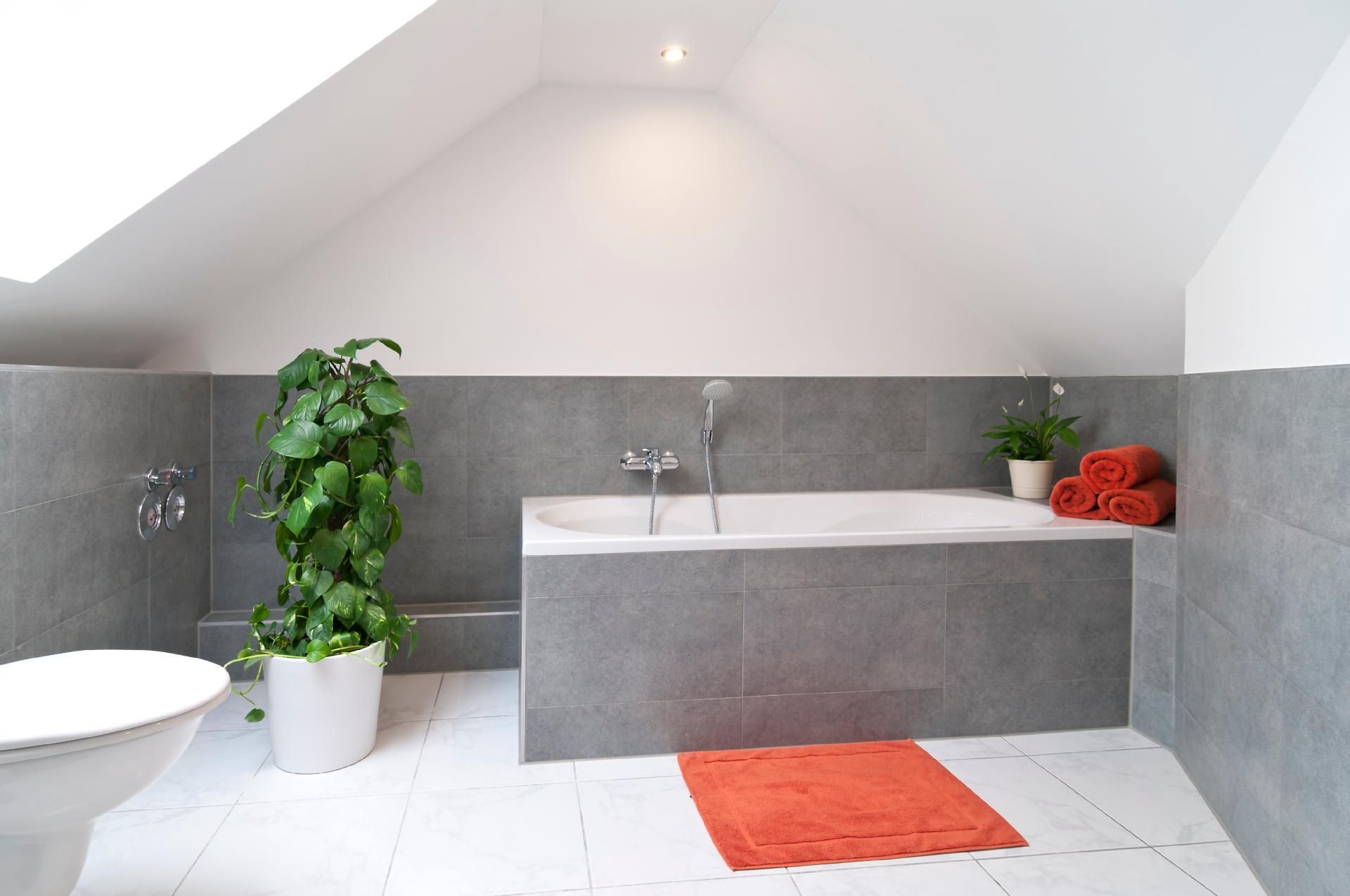 Badezimmer mit Badewanne, grauer halbhoch gefliester Wand und orangefarbenen Handtüchern auf einem weiß gefliesten Boden.