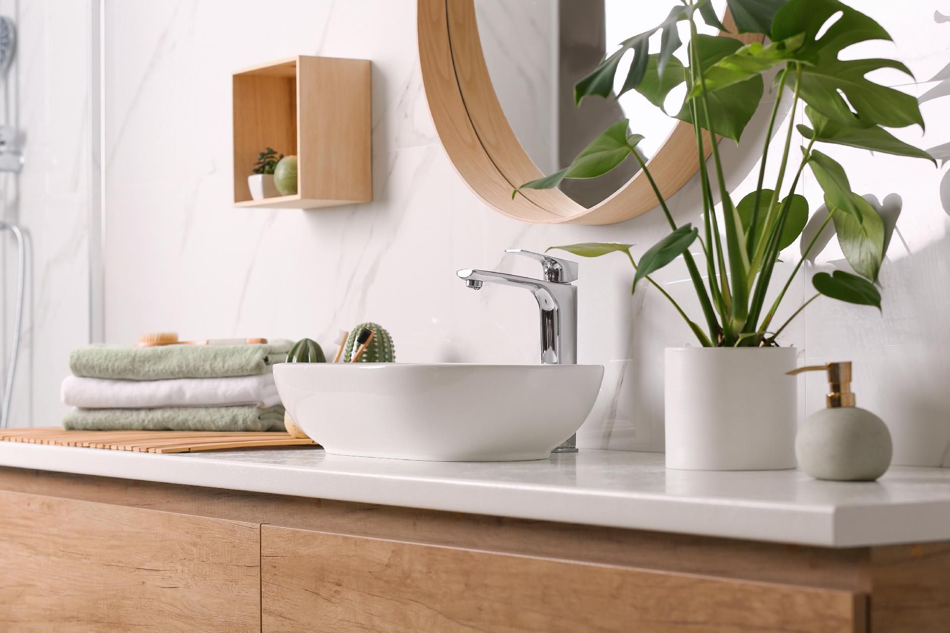 Ein moderner Badezimmerschrank mit Aufsatzwaschbecken, Holzschrank, Handtüchern, Pflanze und rundem Spiegel an der Wand.