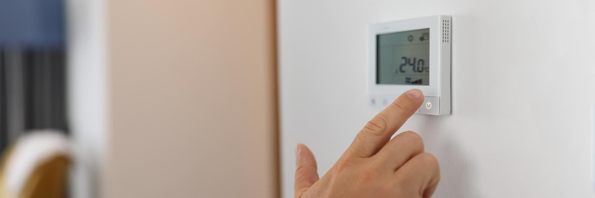 Eine Hand berührt einen weißen digitalen Thermostat an der Wand, der eine Temperatur von 24,0 anzeigt.