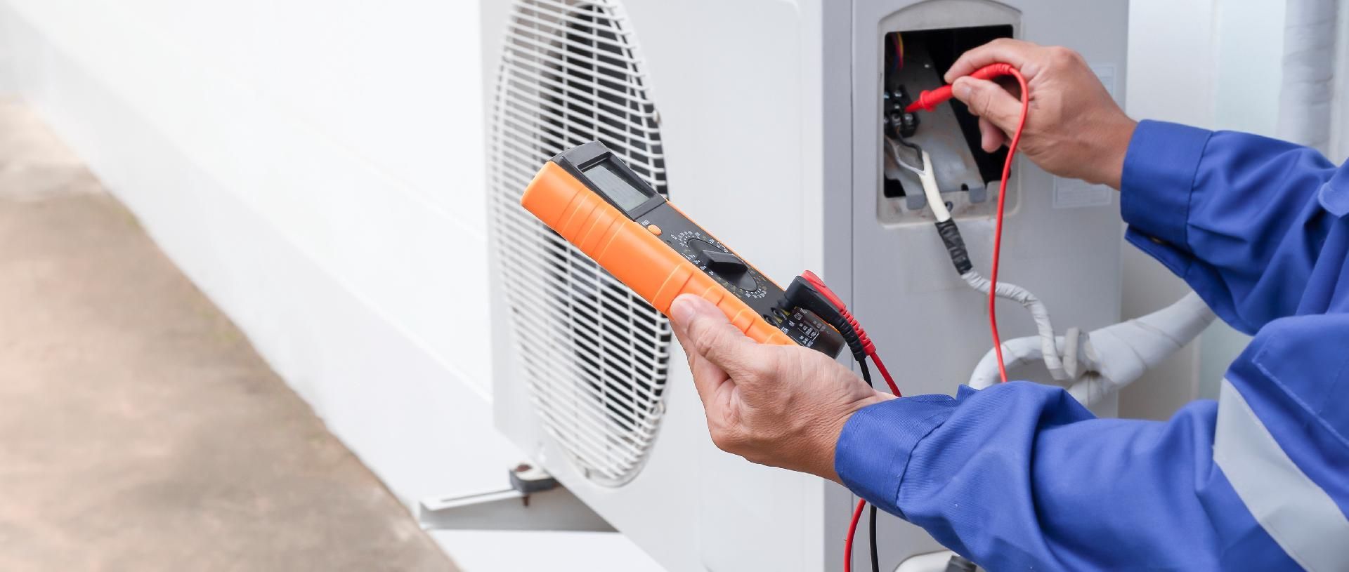 Elektriker in blauer Arbeitskleidung prüft mit einem Multimeter die Drähte einer Außeneinheit einer Klimaanlage.