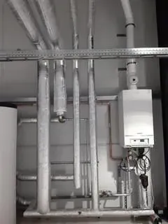 Ein an der Wand montierter Gaskessel, der über isolierte, vertikale Rohre an einer weißen Wand angeschlossen ist.