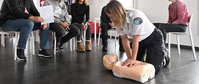 Un groupe de personnes est assis autour d'une ambulancière qui montre les 1ᵉʳˢ secours sur un mannequin