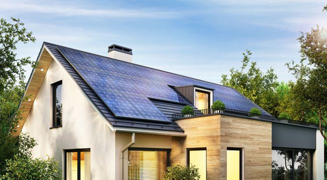 Hände halten ein Tablet, auf dem eine Smart-Home-Steuerungsoberfläche angezeigt wird; im Hintergrund ein weißer Tisch.