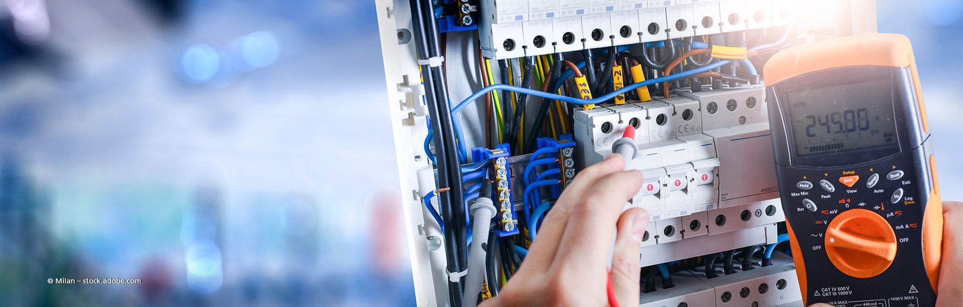Ein Elektriker arbeitet an einem Sicherungskasten und hält ein kleines Werkzeug in der Hand. Rote und blaue Drähte sind sichtbar.