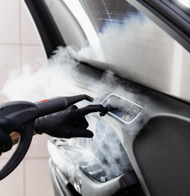 Una mano enguantada limpia el interior de un coche con un limpiador a vapor. El vapor envuelve los controles del salpicadero.