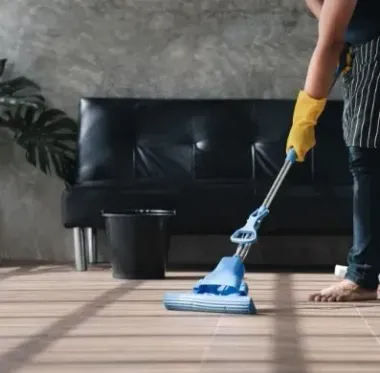 Persona trapeando un piso de madera con un trapeador azul, con guantes y delantal, al lado de un sofá negro y un balde.