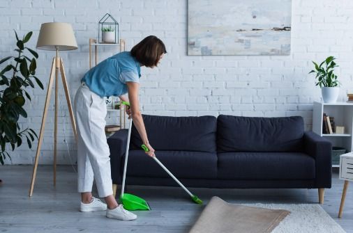 Mujer barriendo una alfombra en una sala de estar, vestida con pantalones blancos y una camisa azul, al lado de un sofá oscuro.
