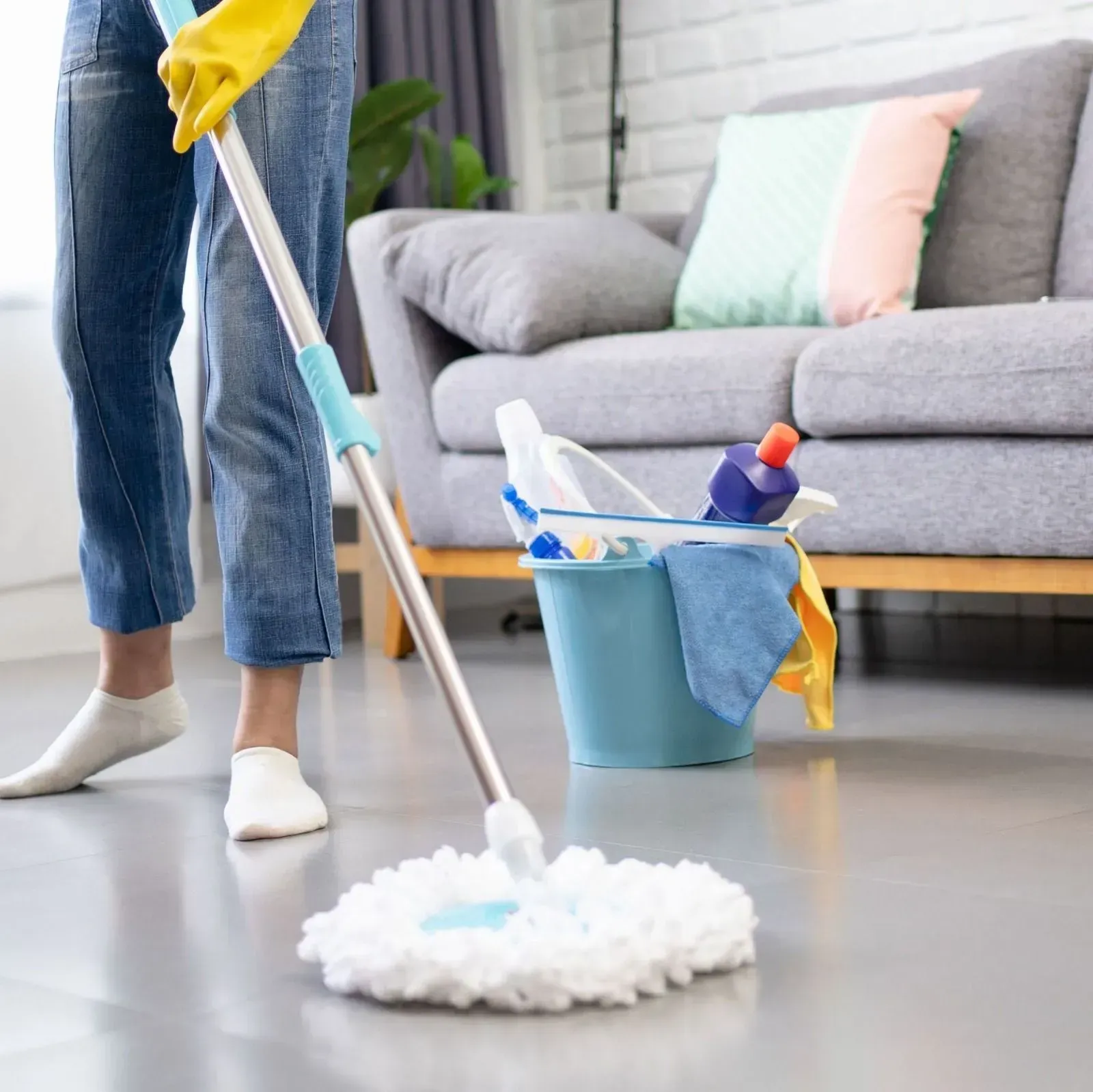 Persona fregando el suelo. Hay guantes amarillos, vaqueros y un cubo de limpieza con suministros.
