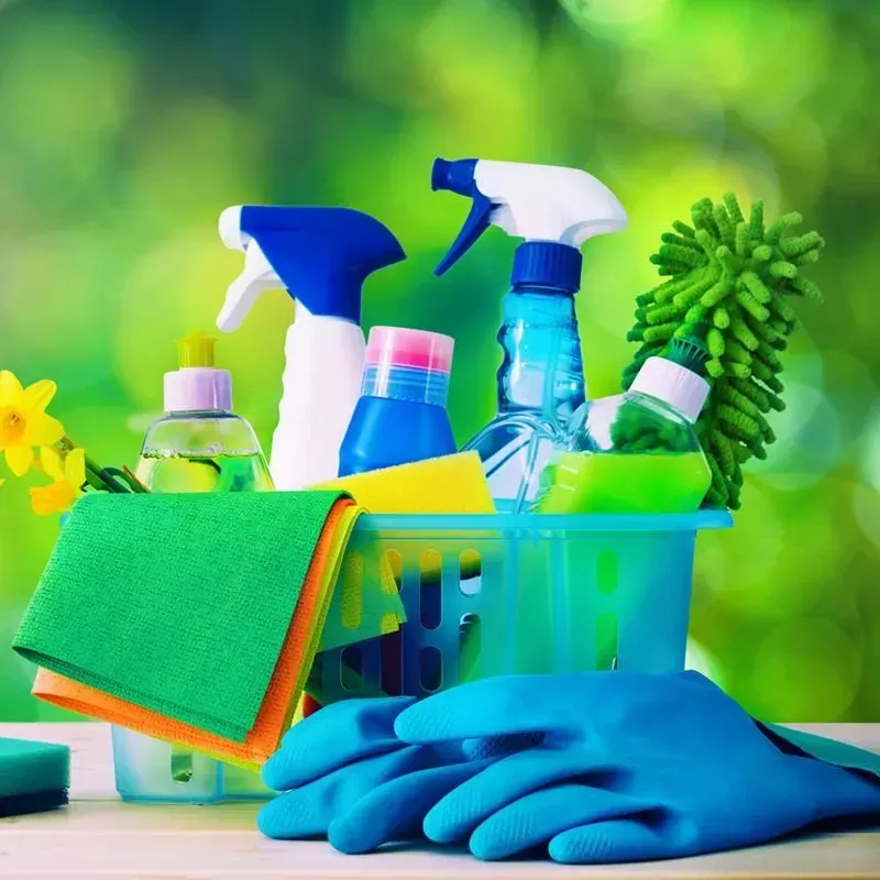 Artículos de limpieza en una cesta con guantes azules, paños verdes y naranjas y botellas de spray sobre un fondo verde borroso.