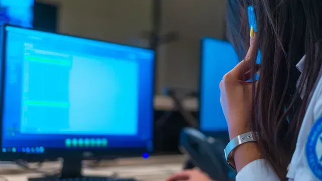 Una mujer con uniforme blanco y emblema azul mira la pantalla de una computadora mientras sostiene un bolígrafo. La pantalla muestra contenido azul.