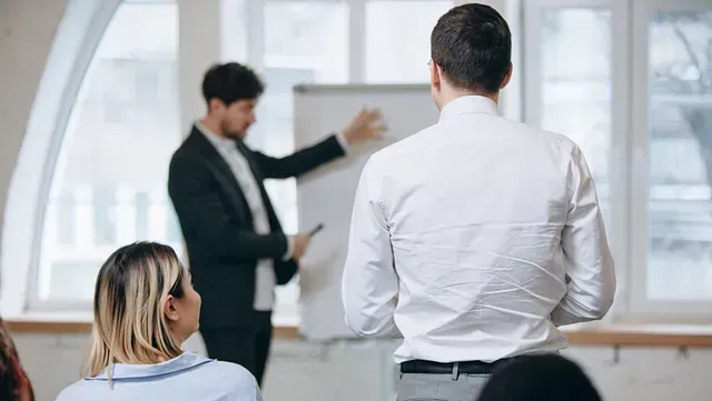 Hombre dando una presentación en una pizarra, visto desde atrás. El público escucha atentamente en una sala iluminada.